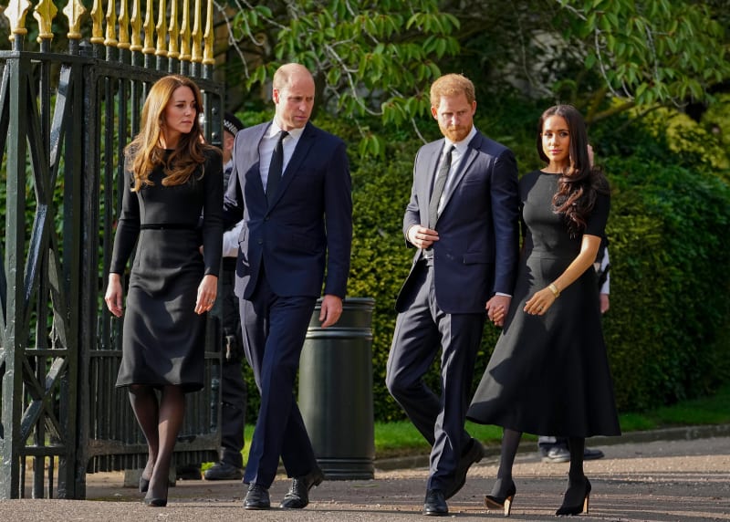 Kate Middleton, Prince William, Prince Harry and Meghan Markle in formal black and dark suits walking together past an ornate black gate