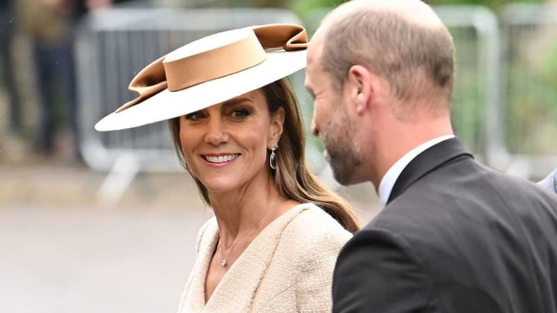 Kate Middleton in a cream outfit and tan-and-white hat smiles at Prince William in a dark suit at an outdoor formal event.