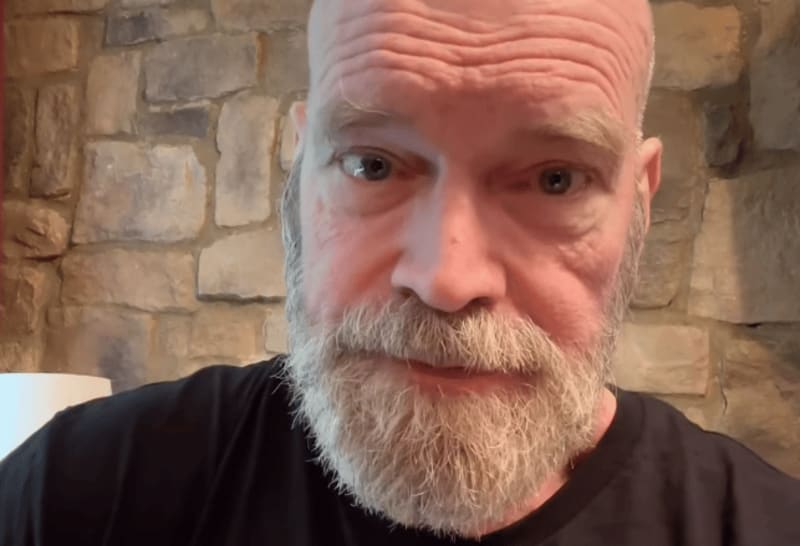 Tyler Mane with a full gray beard wearing a black shirt, photographed against a brick wall background.