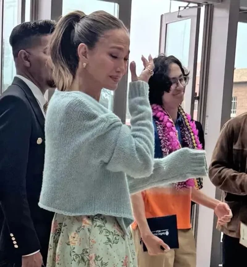 Jennifer Lopez in a light blue sweater and floral skirt wiping away tears at a graduation ceremony.