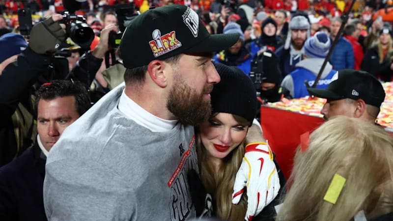 A man in a grey hoodie and black Super Bowl cap embraces a woman in a black beanie from behind, surrounded by a crowd and camera flashes on a football field.