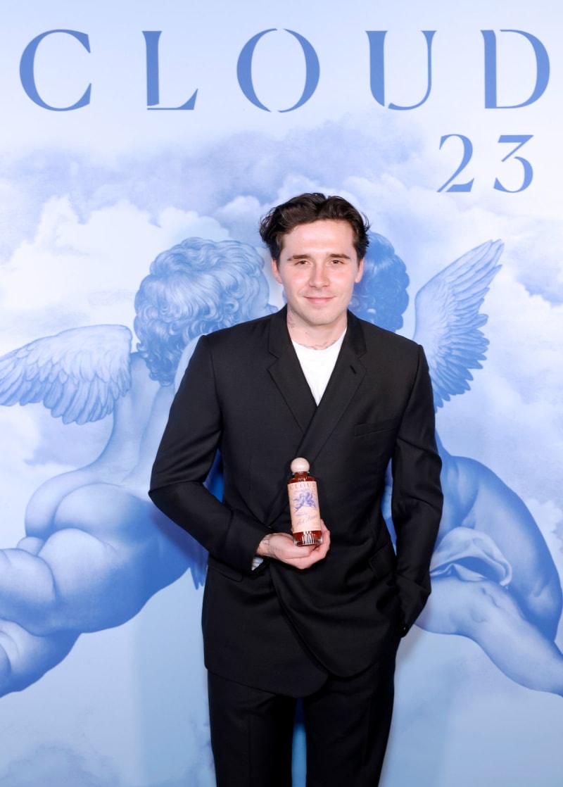 Brooklyn Beckham in a black suit holds a bottle at a "Cloud 23" event backdrop featuring angelic imagery and clouds.