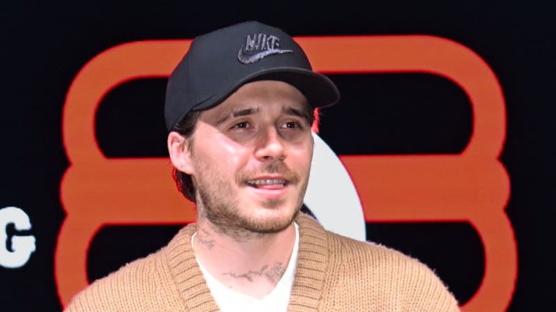 Brooklyn Beckham wearing a black Nike baseball cap and tan cardigan smiles against an orange geometric background.