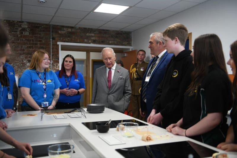 King Charles in a gray suit speaks with young staff members in blue uniforms during a kitchen facility tour.