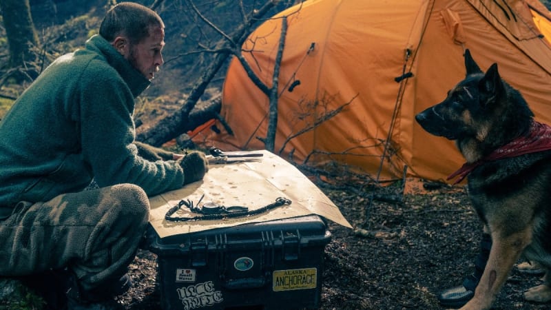 Brad Pitt in a green jacket sits by a campfire with his dog, examining gear near an orange tent and equipment case.