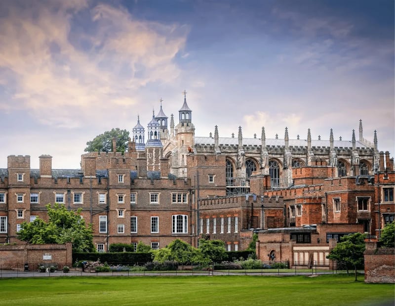 Eton College's historic red brick buildings and Gothic chapel towers against a cloudy sky, viewed from the playing fields.