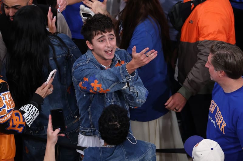 A man in a denim jacket with orange and blue graphic patches, surrounded by fans and crowd at a basketball arena.