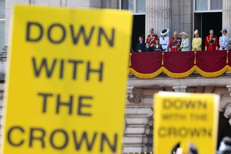 "Down With The Crown" protest placards in the foreground with the Royal Family visible on the Buckingham Palace balcony during Trooping the Colour.