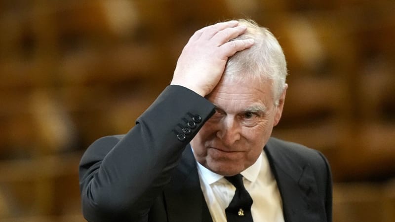 An older white-haired man in a dark suit with a black tie raises his right hand to the top of his head, standing in front of a blurred wooden interior backdrop.