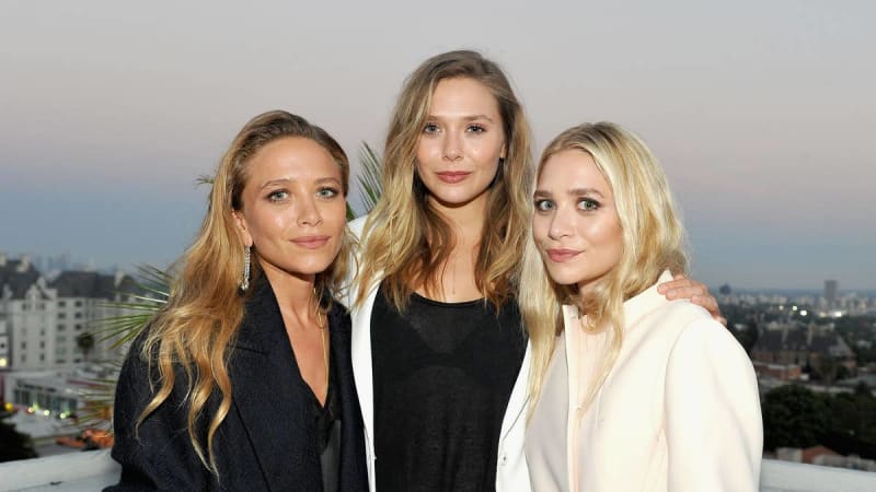 Mary-Kate, Elizabeth and Ashley Olsen with blonde hair pose together outdoors at dusk with a cityscape visible in the background.