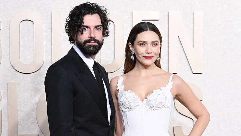 Robbie Arnett and Elizabeth Olsen formal event; Arnett in black suit and tie, Olsen in white lace dress with red lipstick against a cream backdrop.