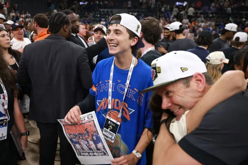 Timothée Chalamet wearing a New York Knicks jersey and cap, holding a newspaper with the headline "Champs!" amid a crowd.