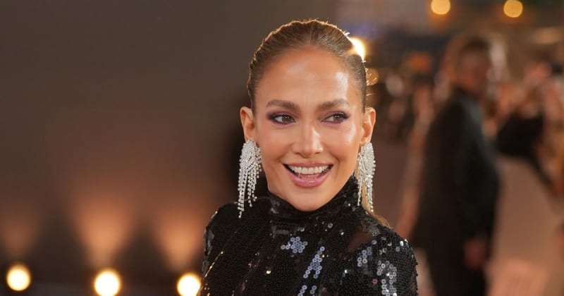 Jennifer Lopez wearing a black sequined outfit and dangling pearl earrings smiles at a formal event with blurred lights in the background.