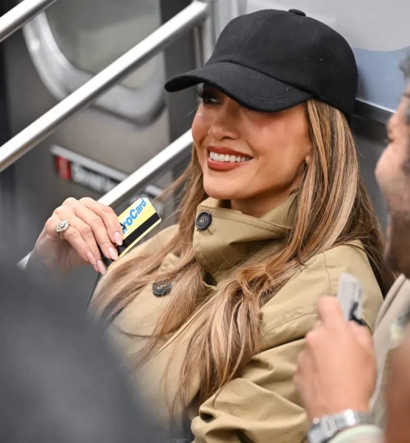 Jennifer Lopez wearing a black cap and tan jacket, smiling while holding a Metro card on a subway train.