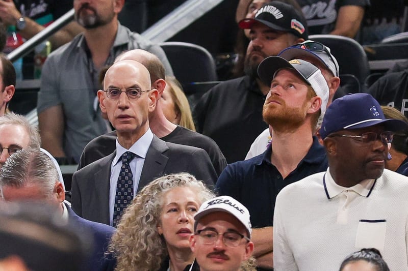 Adam Silver in a suit and tie, and Prince Harry wearing a baseball cap and navy polo shirt, seated among a crowd at a Knicks basketball game.