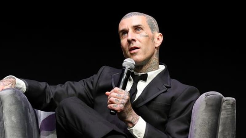 Travis Barker in a black tuxedo with a bow tie holds a microphone while seated, speaking at what appears to be a formal event.