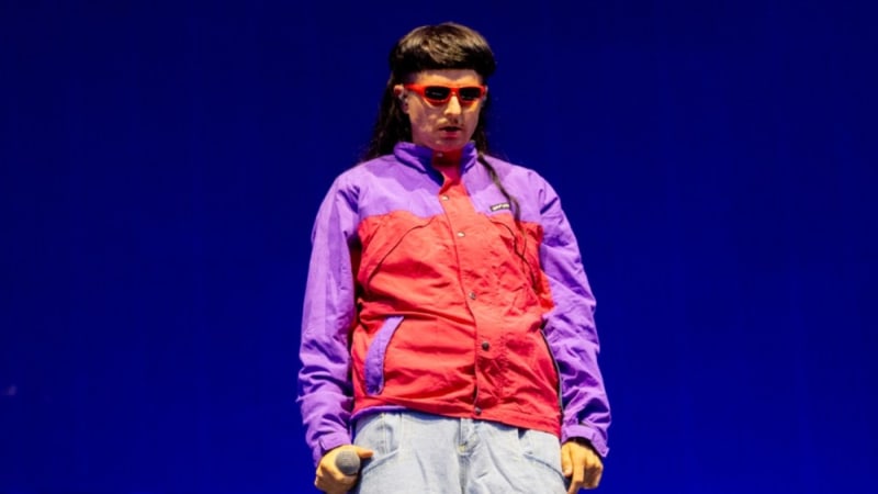 Oliver Tree wearing a colorful red and purple windbreaker, yellow gloves, and red-tinted sunglasses stands against a blue background.
