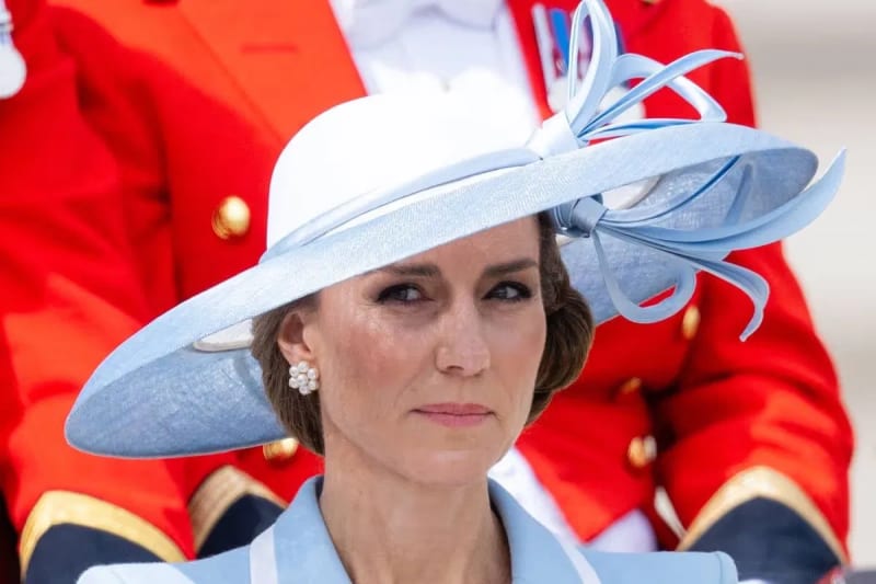 Kate Middleton wearing a pale blue wide-brimmed hat with ribbon detail and pearl earrings, with guardsmen in red uniforms in the background.