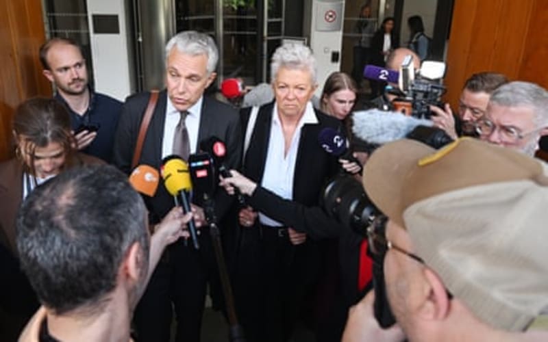 Two prosecutors, a man in a suit and tie and a woman in a black blazer, speaking to a crowd of reporters holding microphones and cameras outside a building.