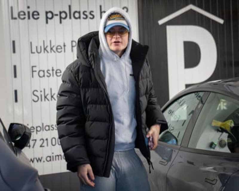 Marius Borg Høiby wearing a black puffer jacket, white hoodie, and cap stands next to a gray car in front of a white building with text.