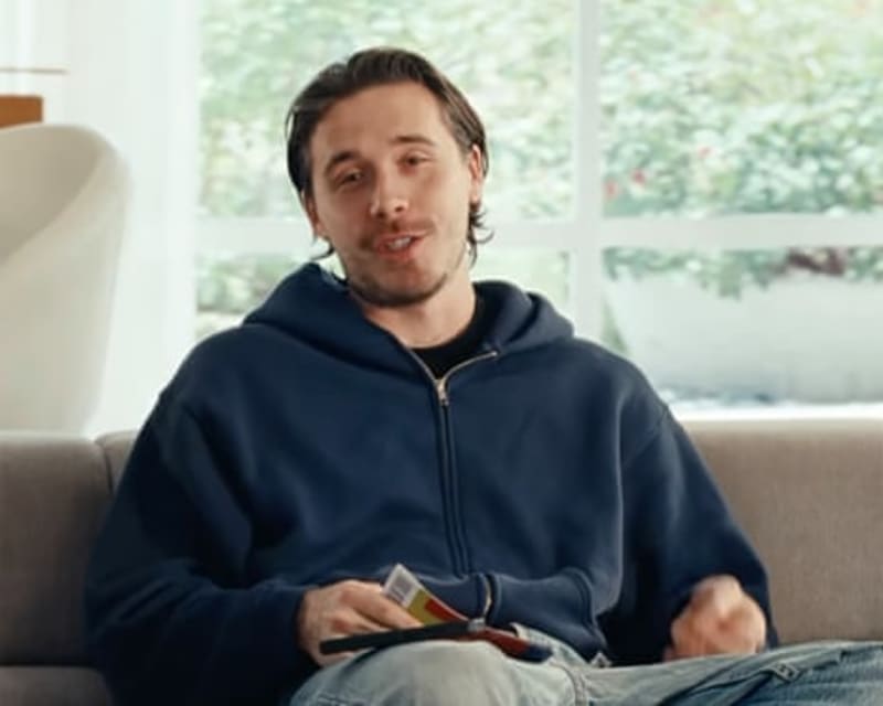 Brooklyn Beckham in a dark blue hoodie sits on a gray couch holding a remote control, with windows behind him.