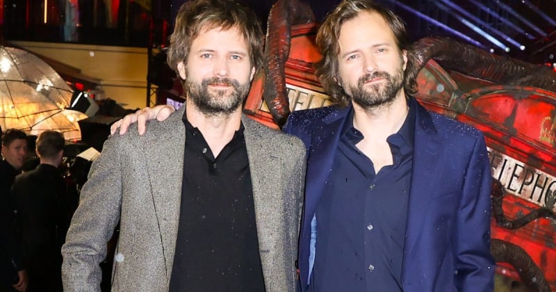 Matt and Ross Duffer in formal attire pose together at an indoor event with red lighting and decorative elements in the background.