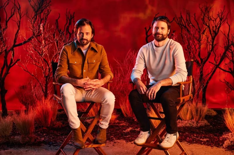 Matt and Ross Duffer seated in director's chairs against a red-lit backdrop with bare black trees, one in a brown shirt jacket and white pants, the other in a cream sweater and dark pants.