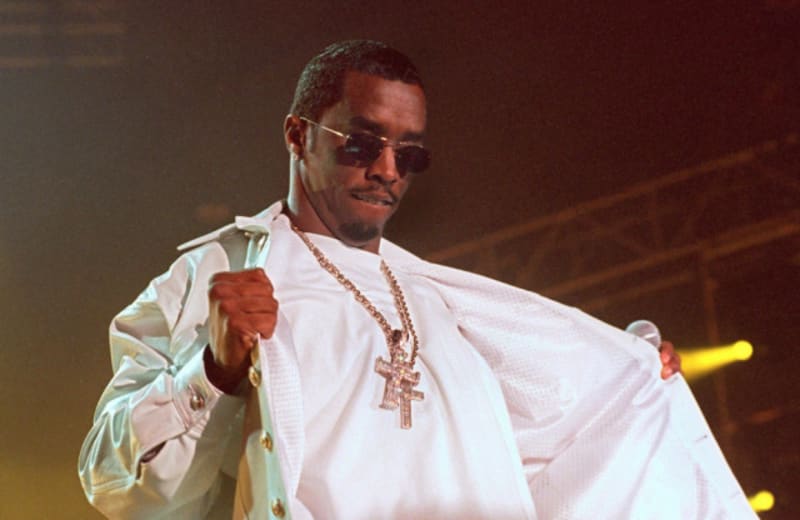 Diddy in sunglasses and white outfit with a diamond cross necklace performs on stage under warm lighting.