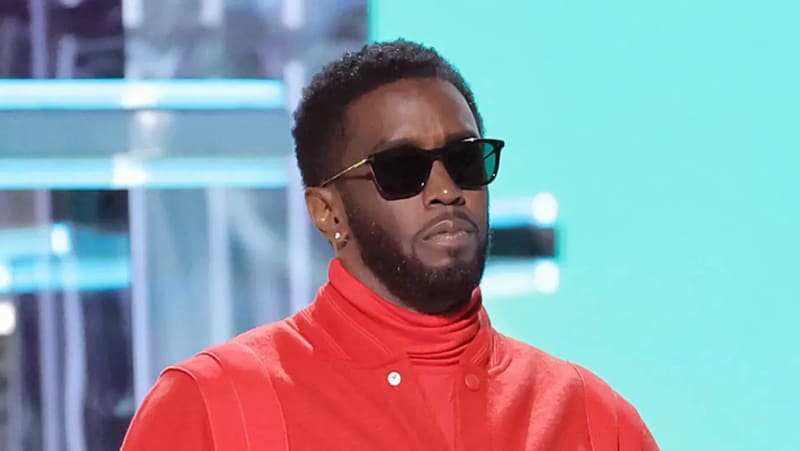 Sean "Diddy" Combs wearing dark sunglasses and a red turtleneck jacket on a stage with a teal backdrop.