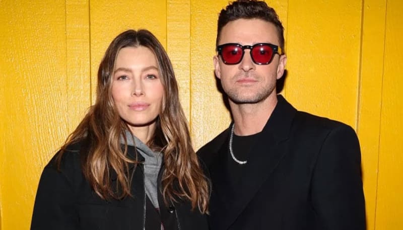 Jessica Biel and Justin Timberlake standing together in front of a yellow wall, both in black outfits, he wearing red-tinted sunglasses and a silver chain necklace.