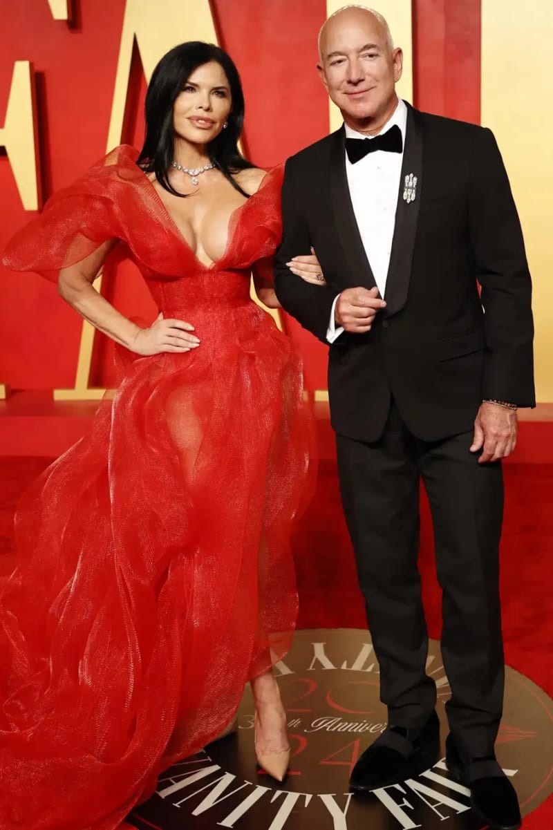Lauren Sanchez in a red ruffled deep-V gown with a diamond necklace, and Jeff Bezos in a black tuxedo, posing together at the Vanity Fair Oscar Party.