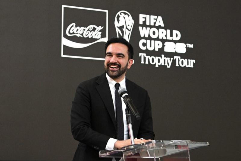 NYC Mayor Zohran Mamdani in a black suit speaking at a podium during a Coca-Cola FIFA World Cup Trophy Tour event.