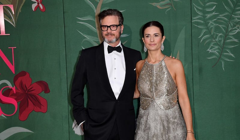 Two people pose together at a formal event in front of a green floral backdrop, one wearing a black tuxedo and the other in a silver sequined dress.
