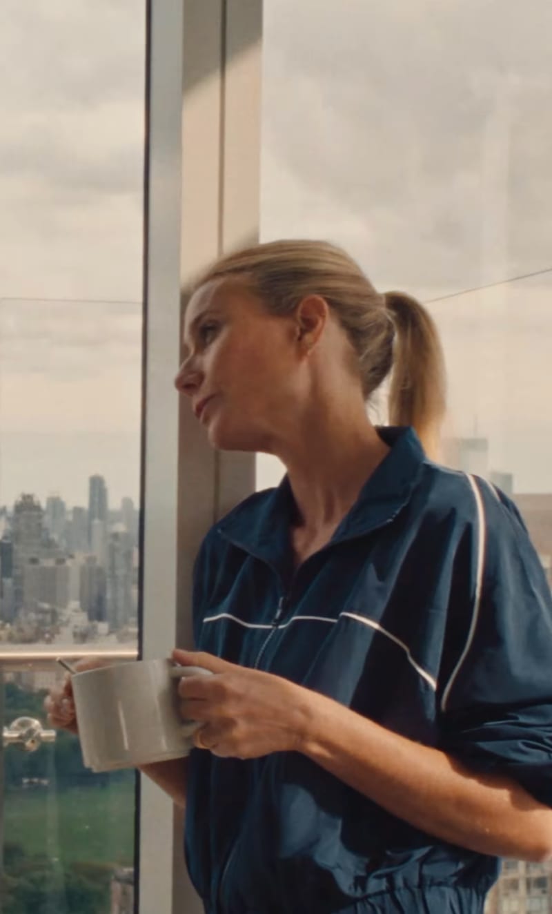 Gwyneth Paltrow in a dark blue shirt holds a white mug while gazing out a high-rise window at a cityscape below.