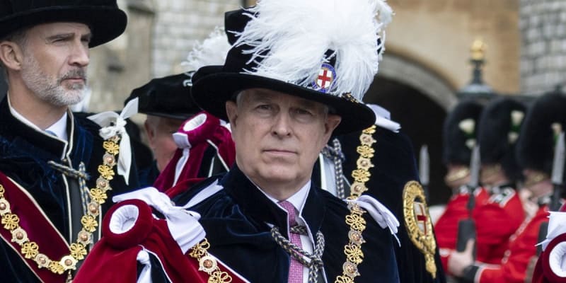 Prince Andrew in formal ceremonial regalia featuring a black hat with white plumes, ornate gold trim, and red velvet robes stands solemnly during an official state occasion.