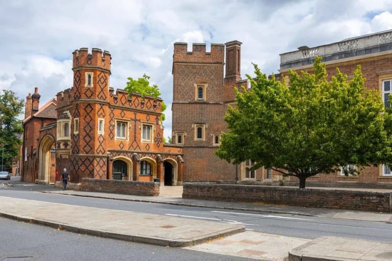 Exterior view of Eton College's red brick buildings with decorative diamond patterning, crenellated towers, and arched entranceways along a quiet street lined with trees.