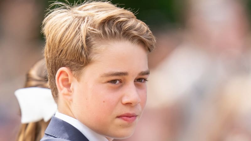 Prince George in formal attire with a white collar and dark jacket, looking to the side with a serious expression.