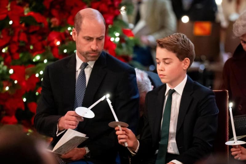 Prince William and Prince George in dark suits holding candles at a Christmas carol service, with poinsettias and a decorated tree in the background.