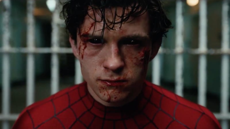Tom Holland as Spider-Man with blood on his face stands in front of a chain-link fence.