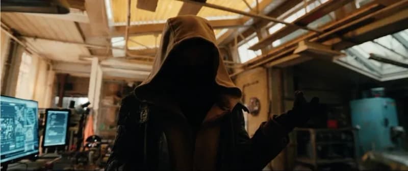 A hooded figure with face obscured standing in a dimly lit industrial space with computer monitors in the background, in a still from Spiderman Brand New Day.