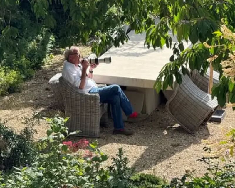 Jeremy Clarkson in a white shirt and jeans sits in a wicker chair photographing in a shaded garden area surrounded by plants.