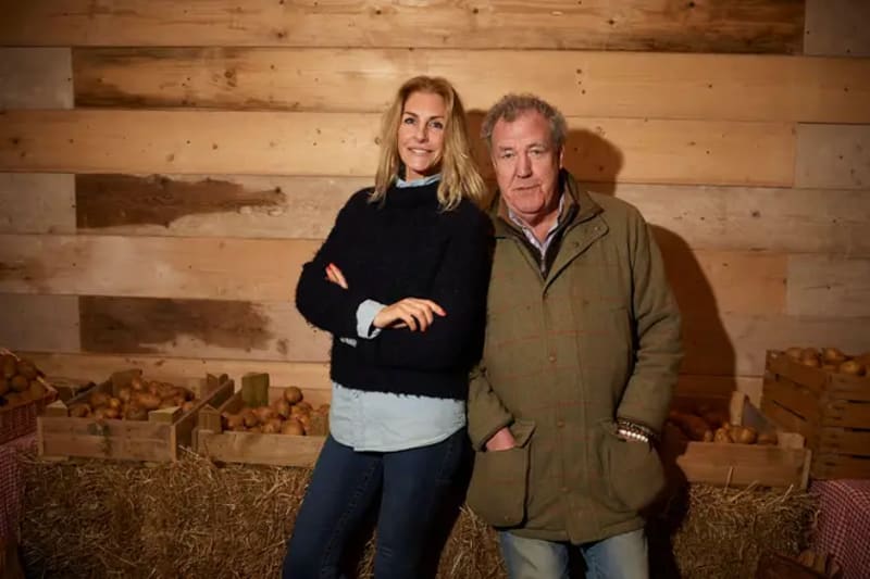 Lisa Hogan and Jeremy Clarkson posing together in front of a wooden wall with hay bales visible in the background.