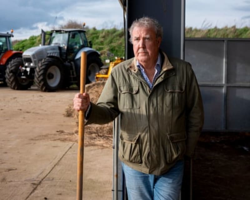 Jeremy Clarkson in olive jacket holds a shovel while standing beside farm equipment and a tractor in a rural setting.