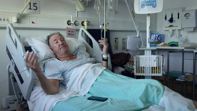 Jeremy Clarkson lies in a hospital bed holding a tablet, wearing a patient gown with medical equipment nearby.