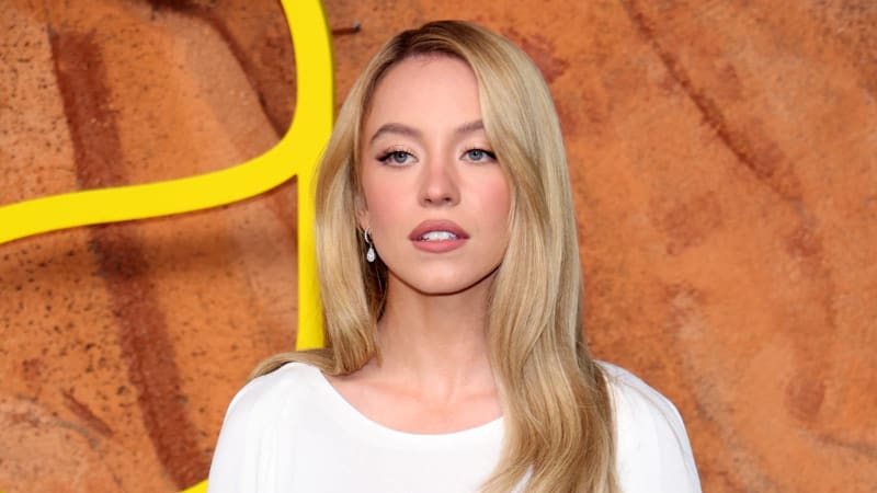 Sydney Sweeney with blonde hair wearing a white top stands against an orange textured backdrop with yellow curved design elements.
