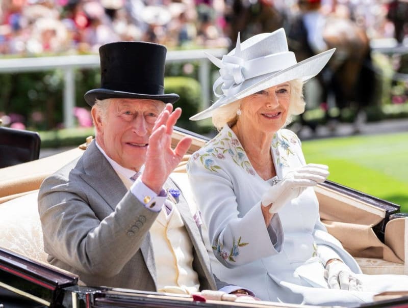 King Charles and Queen Camilla in formal attire wave from an open carriage at what appears to be a royal event or parade.