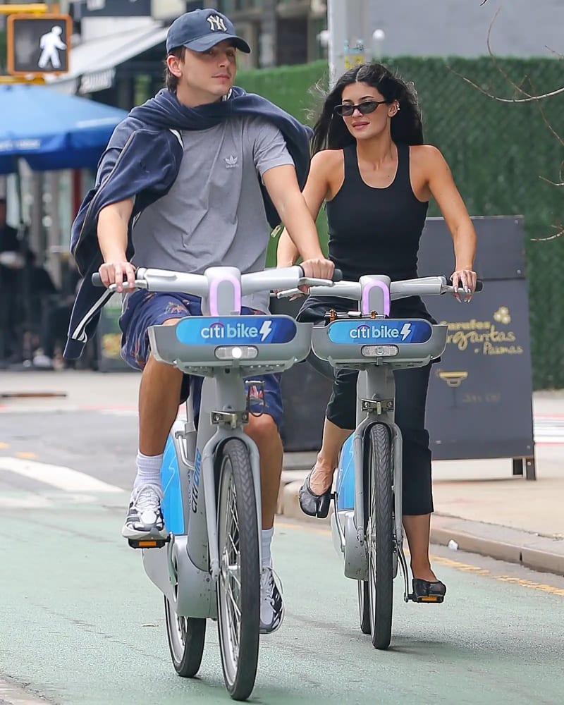 Timothée Chalamet in a grey Adidas t-shirt, navy shorts and Yankees cap, and Kylie Jenner in a black tank top and leggings, riding Citi Bikes side by side on a New York City street.