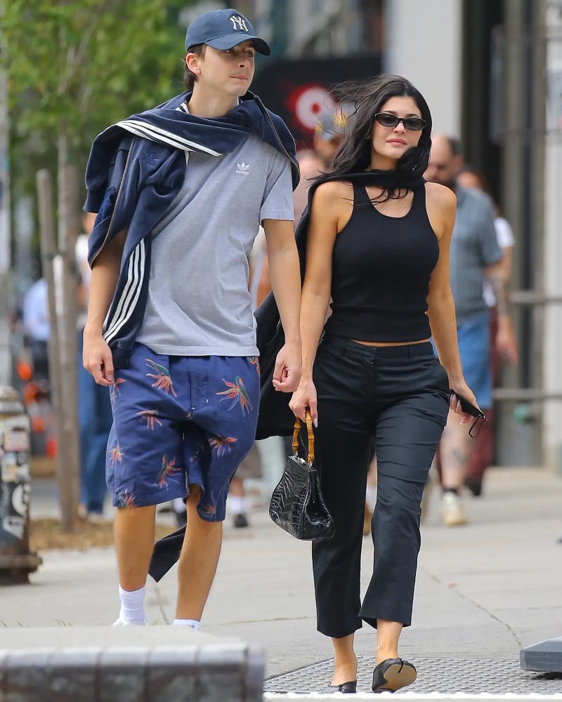 Timothée Chalamet in a grey Adidas t-shirt, floral navy shorts and Yankees cap, and Kylie Jenner in a black cutout tank top, black trousers and carrying a black bamboo-handled handbag, walking together on a New York City sidewalk.