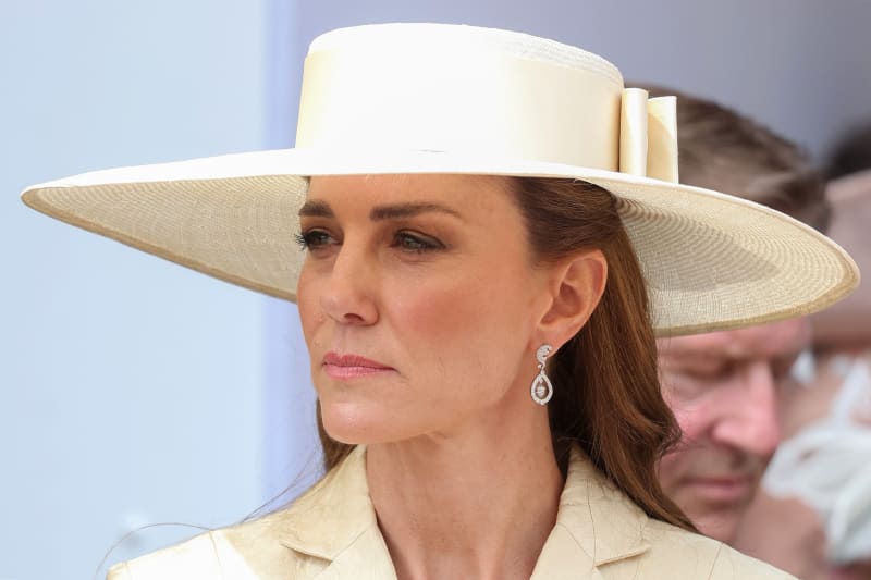 A woman wearing an elegant cream-colored wide-brimmed hat and matching outfit with diamond drop earrings at a formal event.