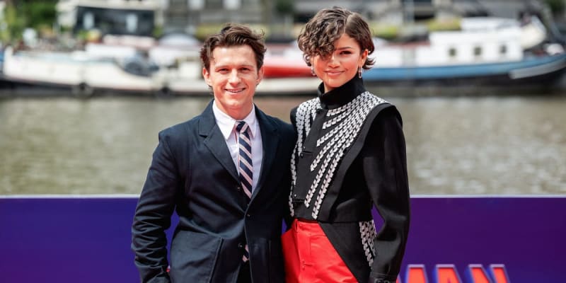 Tom Holland and Zendaya stand together smiling at a waterfront event, with boats visible in the background behind a purple barrier.